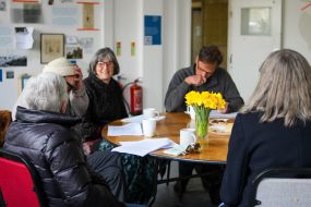 A group of people sat around a table with coffee and some stationary at the shared reading group in St Anne's House